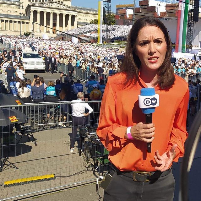Fanny Fascar covering the news by wearing orange shirt and black pant.