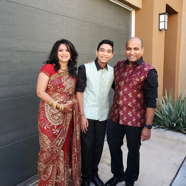 Aryan wearing half coat with black pant and shirt, his mom wearing red designed saree and father red coat with black pant and shirt.