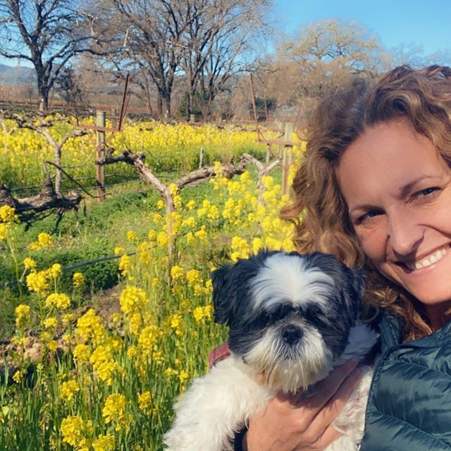 Jerri Manthey posing for the picture in a curly hair and black dress and the flowers on her background with her dog