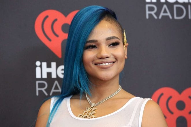 Steph Lecor posing for the photo wearing white tshirt and the locate that has written we the best and with a sparkling blue long hair and smiley face