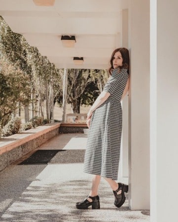 Meghan Heffern wearing black and white dress with black short heels, leaning on the wall